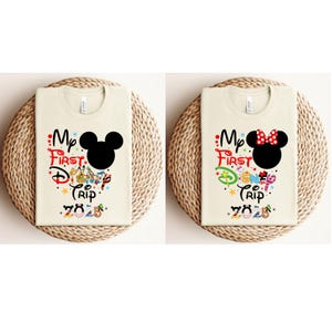 Peut inclure: Deux t-shirts cr&egrave;me avec le texte "My First Disney Trip 2025". L'un pr&eacute;sente une t&ecirc;te de Mickey Mouse noire, l'autre une t&ecirc;te de Minnie Mouse avec un n&oelig;ud rouge. Les chemises sont pli&eacute;es sur un tapis tiss&eacute;.