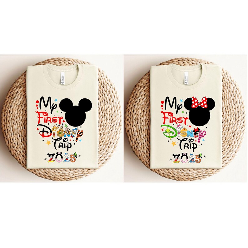 Peut inclure: Deux t-shirts cr&egrave;me avec le texte "My First Disney Trip 2025". L'un pr&eacute;sente une t&ecirc;te de Mickey Mouse noire, l'autre une t&ecirc;te de Minnie Mouse avec un n&oelig;ud rouge. Les chemises sont pli&eacute;es sur un tapis tiss&eacute;.