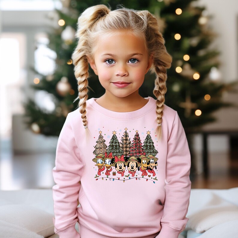 May include: A light pink sweatshirt featuring a festive Christmas design. The graphic includes cartoon Christmas trees and Disney characters like Mickey and Minnie Mouse, Donald Duck, and Goofy, all wearing scarves. The design is accented with string lights.