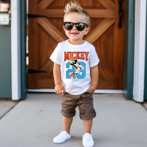 以下が含まれることがあります： 白いTシャツを着た小さな男の子。Tシャツにはミッキーマウスがバスケットボールをしているグラフィックと「MICKEY 23」の文字がプリントされています。彼は茶色のショートパンツ、白いスニーカー、黒いサングラスも着用しています。