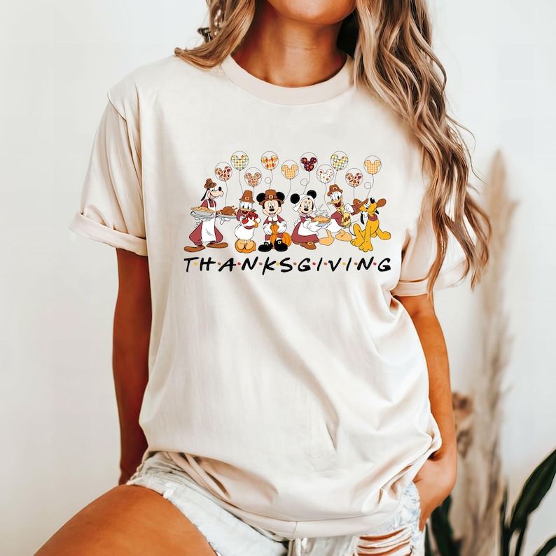 K&ouml;nnte beinhalten: Ein beiges T-Shirt mit einem Aufdruck von Mickey Mouse und seinen Freunden in Thanksgiving-Kost&uuml;men. Der Aufdruck enth&auml;lt den Text "THANKSGIVING" in einer stilisierten Schriftart.