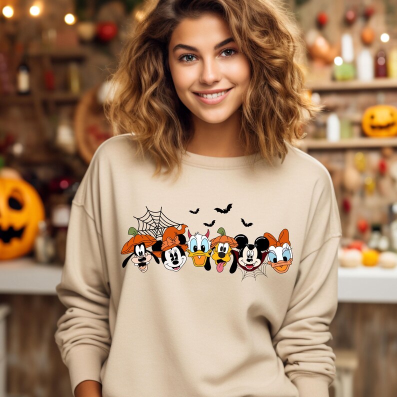 May include: A tan sweatshirt with a graphic of Mickey Mouse, Minnie Mouse, Donald Duck, Goofy, and Pluto dressed in Halloween costumes. The characters are arranged in a row and have a spiderweb and bats behind them.