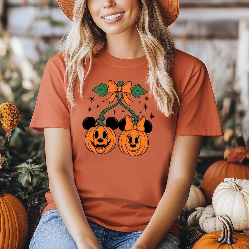 May include: Orange t-shirt featuring a Halloween-themed design. The graphic includes two smiling pumpkin faces with Mickey and Minnie Mouse ears, connected by a green vine and bow. The design is surrounded by stars.