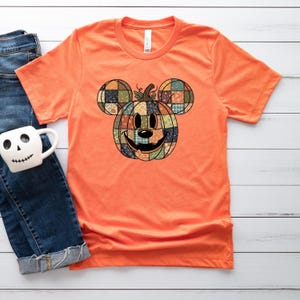 May include: Orange t-shirt featuring a pumpkin design in the shape of a famous mouse. The pumpkin is made of patchwork fabric with a smiling face. The shirt is a short-sleeved crew neck.