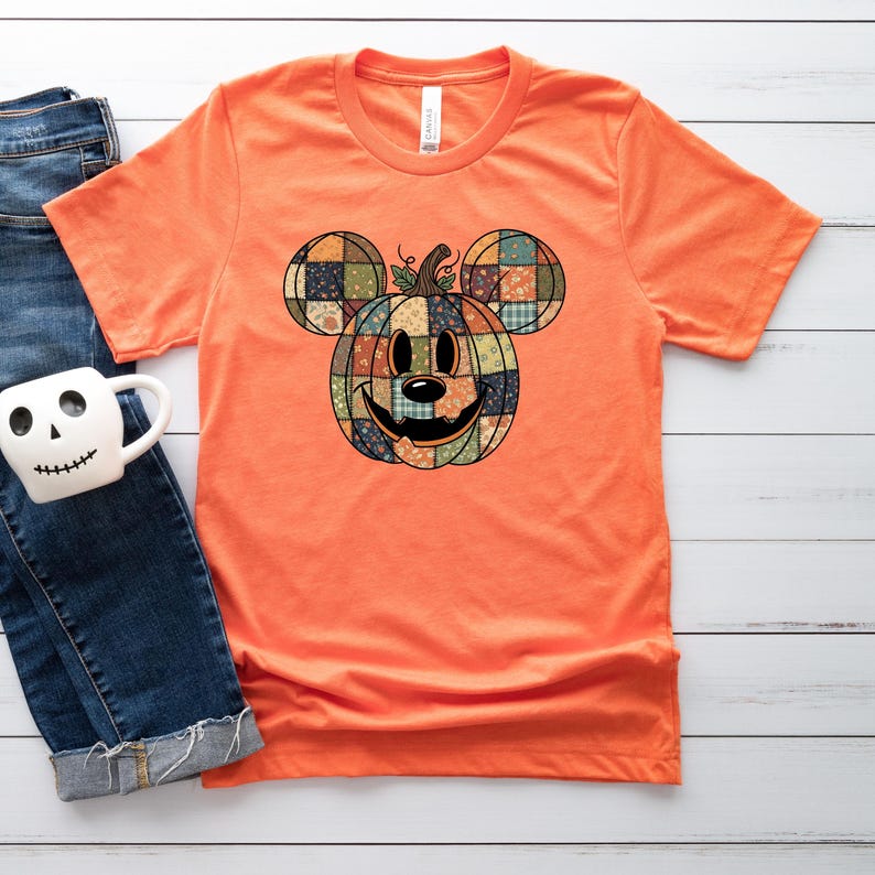 May include: Orange t-shirt featuring a pumpkin design in the shape of a famous mouse. The pumpkin is made of patchwork fabric with a smiling face. The shirt is a short-sleeved crew neck.