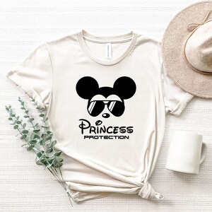 May include: A beige t-shirt with a black and white graphic of Mickey Mouse wearing sunglasses and the text "Princess Protection".