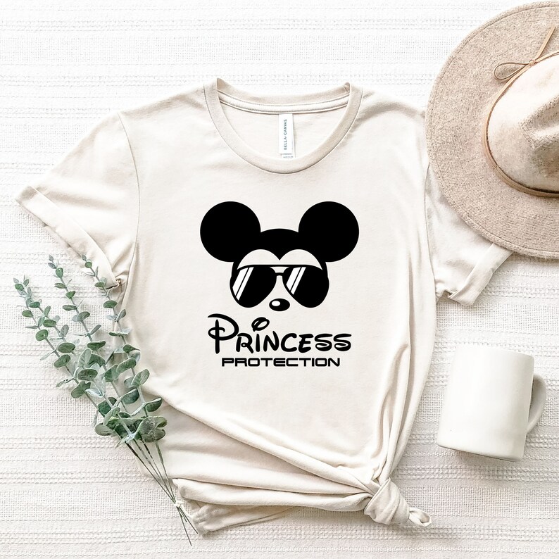 May include: A beige t-shirt with a black and white graphic of Mickey Mouse wearing sunglasses and the text "Princess Protection".