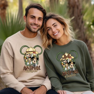 May include: Two sweatshirts with a graphic print of Mickey Mouse and friends in safari hats. The text "Animal Kingdom" is printed below the graphic. One sweatshirt is beige and the other is green.