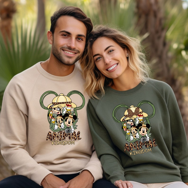 May include: Two sweatshirts with a graphic print of Mickey Mouse and friends in safari hats. The text "Animal Kingdom" is printed below the graphic. One sweatshirt is beige and the other is green.