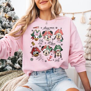 May include: A light pink sweatshirt featuring a graphic of Disney characters dressed as pirates. The characters include Mickey Mouse, Minnie Mouse, Donald Duck, Daisy Duck, Pluto, and Goofy. The words "Mickey Mouse" and character names are also printed.