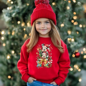 May include: A young person wearing a red sweatshirt and a matching red knit cap. The sweatshirt features a colorful graphic of Mickey and Minnie Mouse in Santa hats, surrounded by Christmas gifts and decorations. The background is a blurred Christmas tree.