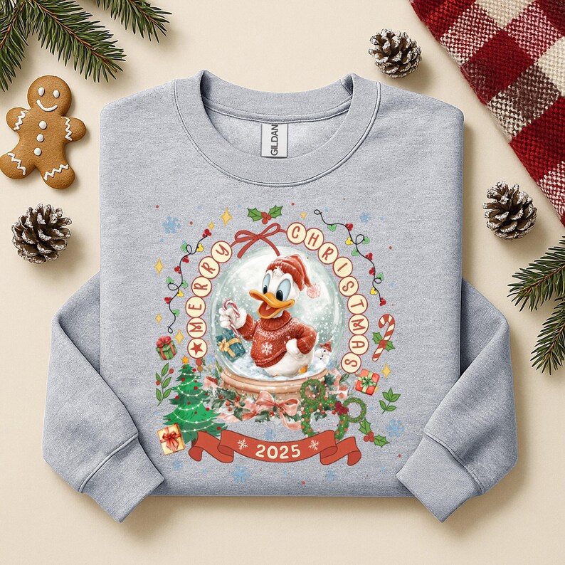 Pode incluir: Camisola cinza claro com um gr&aacute;fico do Pato Donald dentro de uma bola de neve, usando um gorro e su&eacute;ter de Papai Noel. As palavras "MERRY CHRISTMAS" circundam o globo, com "2025" em uma faixa abaixo. Design festivo de f&eacute;rias.