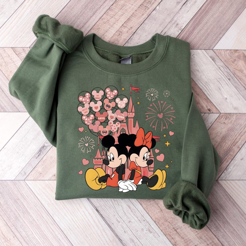 May include: A green sweatshirt with a graphic print of Mickey Mouse and Minnie Mouse in front of a pink castle with balloons and fireworks. The balloons have hearts and the text "LOVE" and "XOXO".