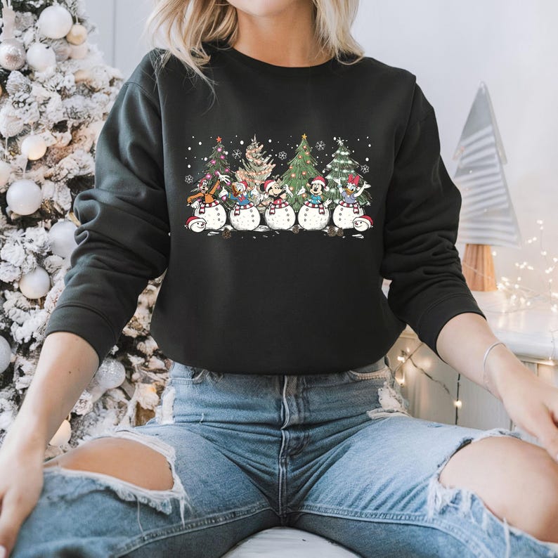 Op de afbeelding: Zwart sweatshirt met een kerstthema ontwerp met Mickey Mouse, Donald Duck, Goofy, Pluto en Minnie Mouse verkleed als sneeuwpoppen. De personages staan voor kerstbomen.