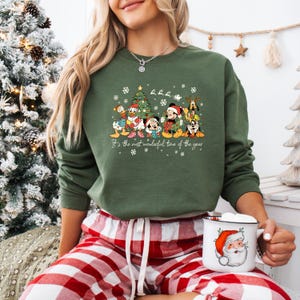 May include: Green crewneck sweatshirt featuring a festive graphic of Disney characters around a Christmas tree. The text "It's the most wonderful time of the year" is printed below the characters. The person is wearing red and white plaid pajama pants and holding a Santa mug.