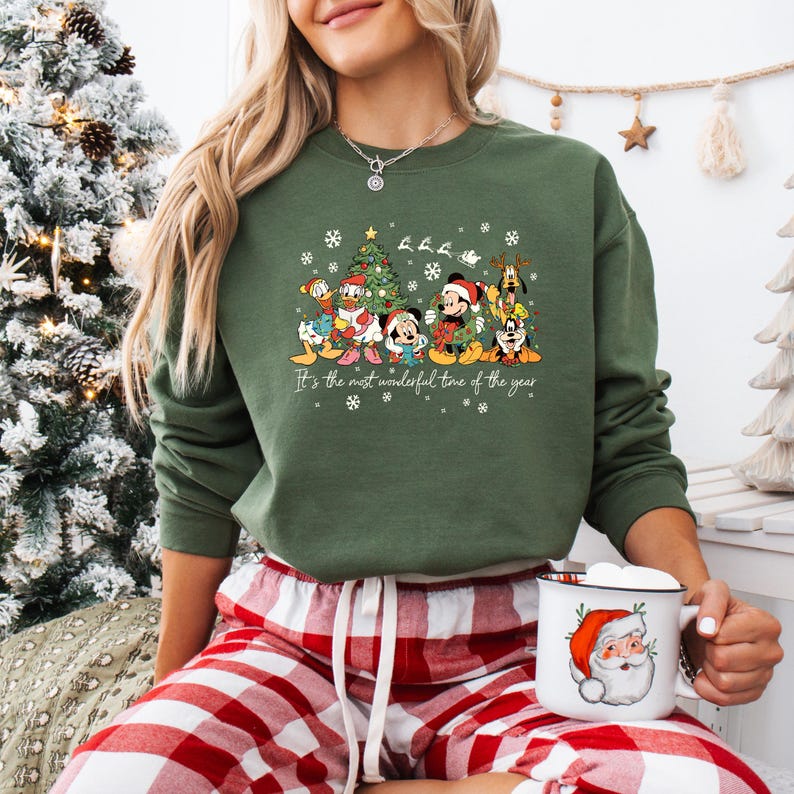 May include: Green crewneck sweatshirt featuring a festive graphic of Disney characters around a Christmas tree. The text "It's the most wonderful time of the year" is printed below the characters. The person is wearing red and white plaid pajama pants and holding a Santa mug.