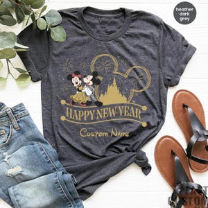 May include: A heather dark grey t-shirt with a gold Mickey Mouse and Minnie Mouse design. The shirt says "Happy New Year" and has a space for a custom name.
