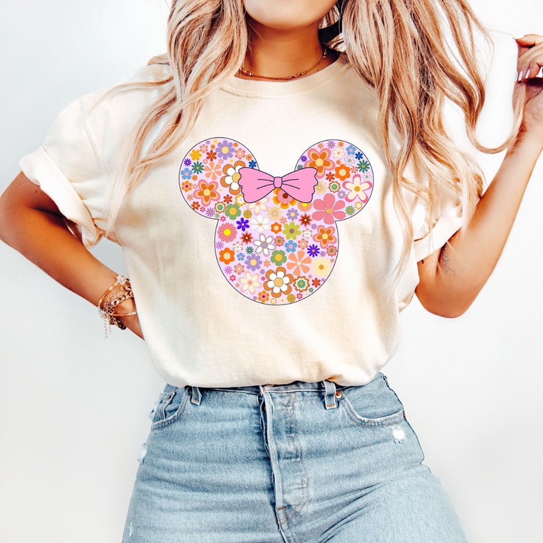 May include: Cream-colored t-shirt featuring a floral Mickey Mouse design. The mouse's head and ears are filled with colorful flowers, and a pink bow sits in the center. The shirt is a casual style.