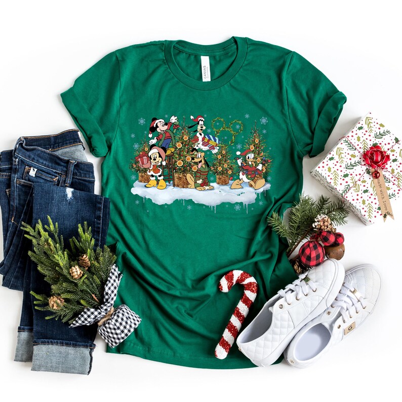 May include: Green t-shirt with a Christmas-themed design featuring Mickey Mouse, Minnie Mouse, Donald Duck, Goofy, and Pluto decorating Christmas trees. The design includes snowflakes and the iconic Disney castle.
