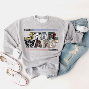 May include: A gray sweatshirt with a graphic of the Star Wars logo. The logo is made up of images of characters from the Star Wars franchise, including Darth Vader, Yoda, and R2-D2.