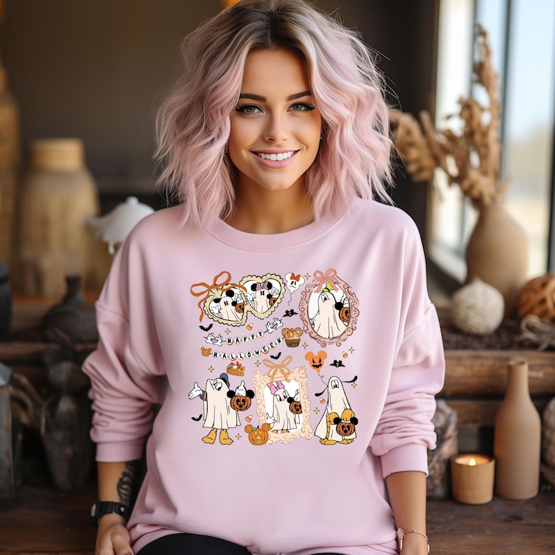 May include: A light pink sweatshirt featuring a Halloween-themed graphic. The graphic includes cartoon ghosts, pumpkins, bats, and the text "Happy Halloween." The design is in shades of white, orange, black, and brown.