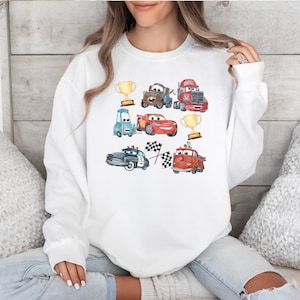 May include: White sweatshirt with a colorful cartoon design featuring various cars, trucks, and trophies. The cars are red, blue, green, and brown. The trucks are red and blue. The trophies are gold.