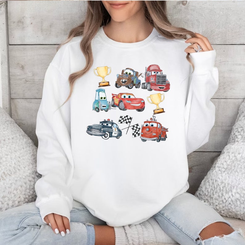 May include: White sweatshirt with a colorful cartoon design featuring various cars, trucks, and trophies. The cars are red, blue, green, and brown. The trucks are red and blue. The trophies are gold.