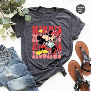 K&ouml;nnte beinhalten: Dunkelgraues T-Shirt mit einem Aufdruck von Mickey und Minnie Mouse, die sich umarmen, mit den Worten "MINNIE MOMMY" in Rot und Rosa. Das Bild zeigt blaue Jeans und braune Sandalen.