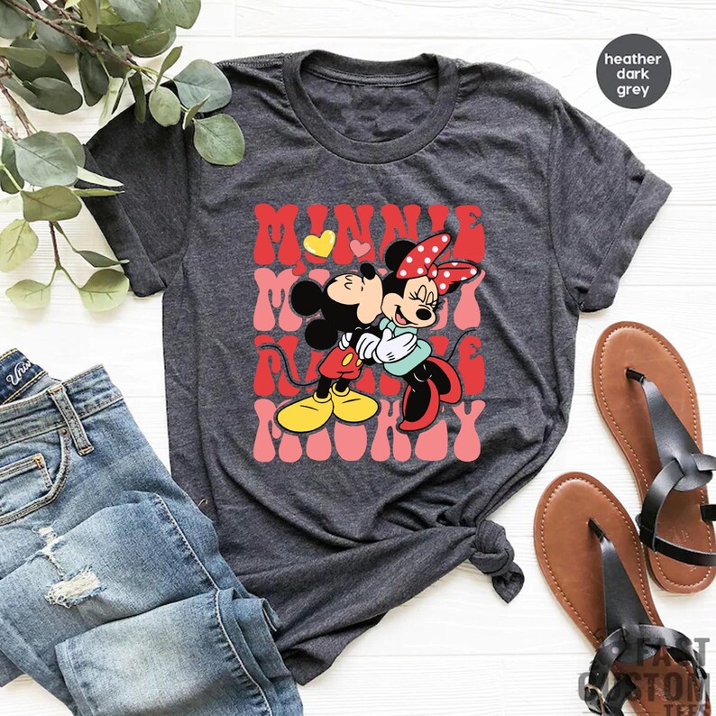 K&ouml;nnte beinhalten: Dunkelgraues T-Shirt mit einem Aufdruck von Mickey und Minnie Mouse, die sich umarmen, mit den Worten "MINNIE MOMMY" in Rot und Rosa. Das Bild zeigt blaue Jeans und braune Sandalen.