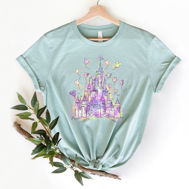 May include: A light blue t-shirt featuring a watercolour illustration of a castle with Mickey Mouse balloons. The castle is outlined in black and filled with shades of purple, pink and yellow.