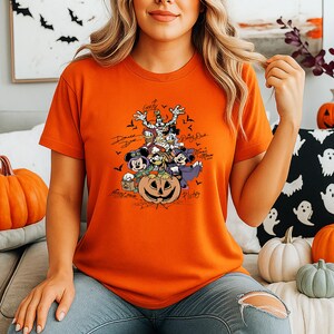 May include: Orange t-shirt featuring a Halloween-themed graphic with Disney characters, including Mickey Mouse, Minnie Mouse, Donald Duck, and Goofy, atop a carved pumpkin. The shirt has character signatures and small bats.