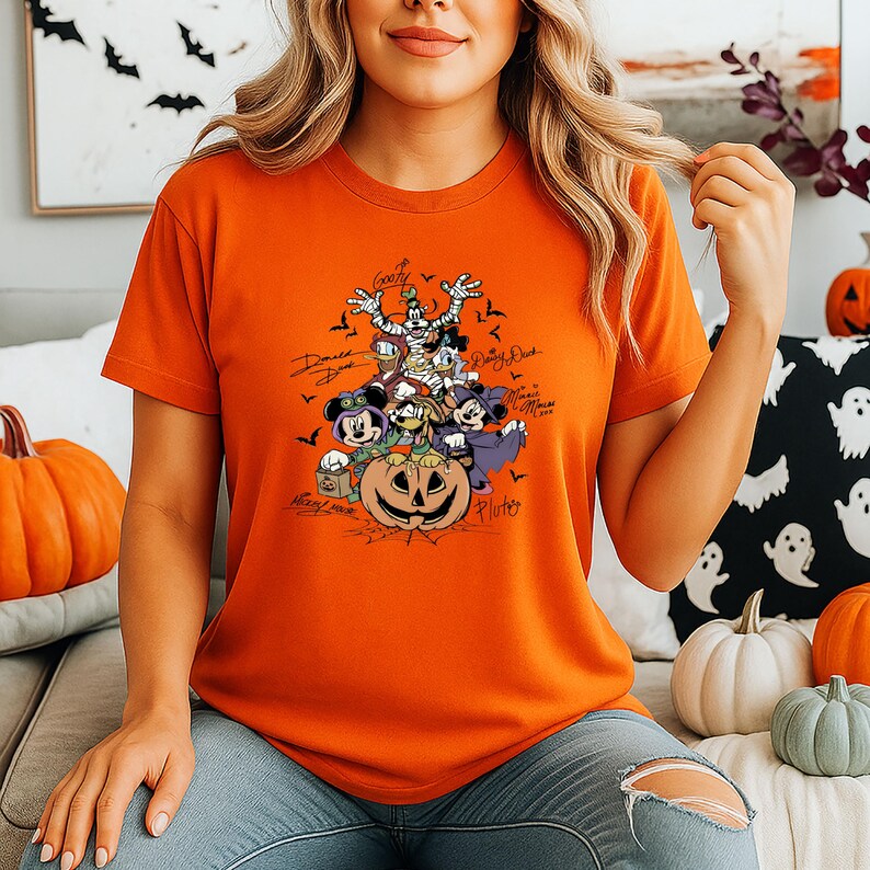 May include: Orange t-shirt featuring a Halloween-themed graphic with Disney characters, including Mickey Mouse, Minnie Mouse, Donald Duck, and Goofy, atop a carved pumpkin. The shirt has character signatures and small bats.