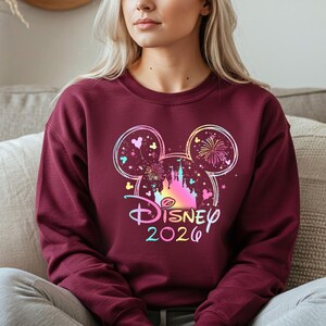 May include: Burgundy crewneck sweatshirt featuring a colorful Disney design. The design includes a castle, fireworks, and Mickey Mouse ears, with the word "Disney" and the year "2020" in a rainbow gradient.