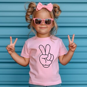 K&ouml;nnte beinhalten: Ein junges Kind tr&auml;gt ein rosa T-Shirt mit einem schwarzen Peace-Zeichen-Motiv. Das Kind tr&auml;gt au&szlig;erdem eine rosa Sonnenbrille und eine rosa Schleife. Das Kind h&auml;lt zwei Finger jeder Hand als Friedenszeichen hoch.