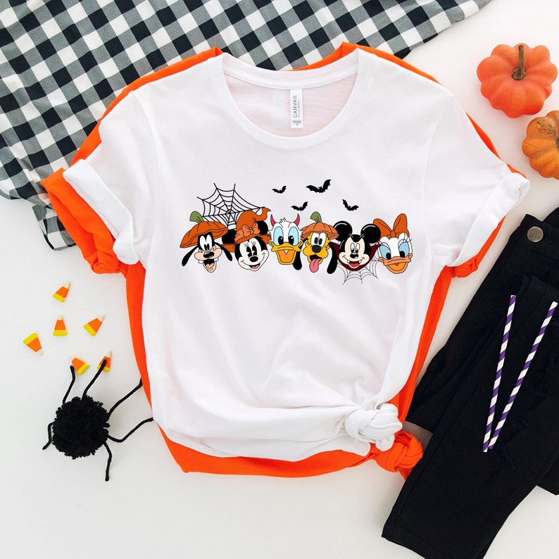 May include: White t-shirt with a graphic of Mickey Mouse, Minnie Mouse, Donald Duck, Goofy, and Pluto dressed in Halloween costumes. The characters are lined up in a row with a spider web and bats above them.
