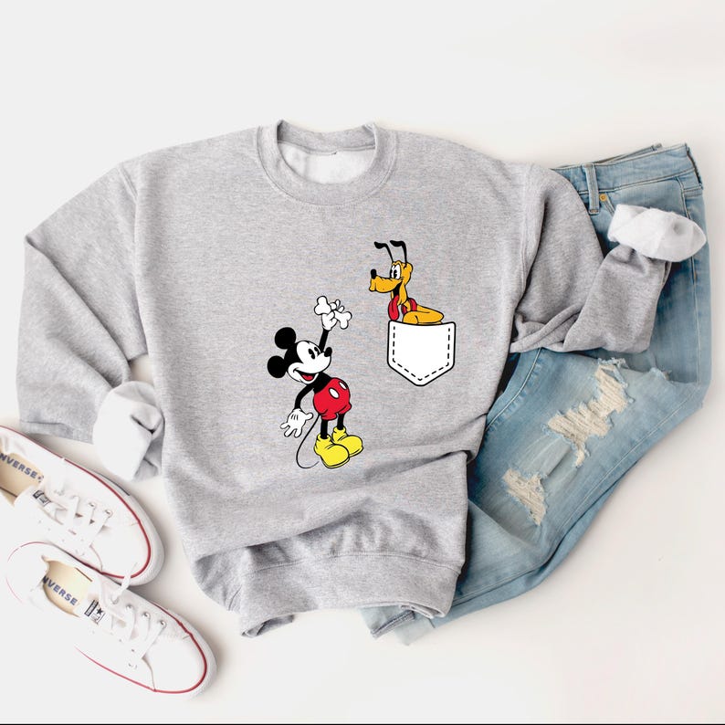 Puede incluir: Una sudadera gris con un bolsillo blanco que presenta una imagen de dibujos animados de Pluto el perro. Mickey Mouse est&aacute; de pie a la izquierda del bolsillo, sosteniendo un hueso.