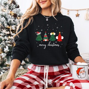 May include: Black crewneck sweatshirt featuring four Christmas tree designs with festive decorations and the words "merry christmas" in white script. The trees are green and red, with snowflakes scattered above.