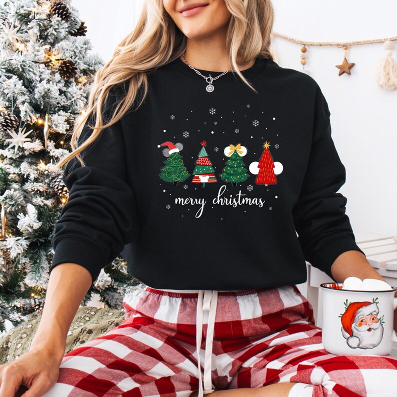 May include: Black crewneck sweatshirt featuring four Christmas tree designs with festive decorations and the words "merry christmas" in white script. The trees are green and red, with snowflakes scattered above.