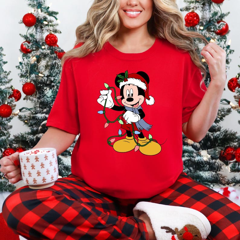 Puede incluir: Una camiseta roja con un personaje de dibujos animados de Mickey Mouse con un gorro de Pap&aacute; Noel y una bufanda, sosteniendo luces navide&ntilde;as. La camiseta forma parte de un atuendo navide&ntilde;o, combinada con un pantal&oacute;n de pijama a cuadros rojos y negros.