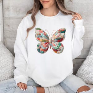 May include: White sweatshirt featuring a colorful butterfly design. The butterfly is made of patchwork fabric in various colors, including red, blue, green, and yellow. The sweatshirt is a casual, comfortable style.
