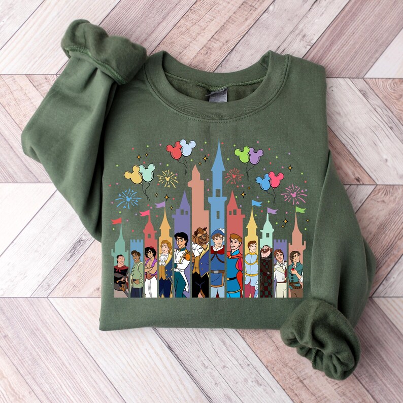 Puede incluir: Sudadera verde con una ilustraci&oacute;n colorida de pr&iacute;ncipes Disney frente a una silueta de castillo. Globos de Mickey Mouse y fuegos artificiales est&aacute;n en el fondo.