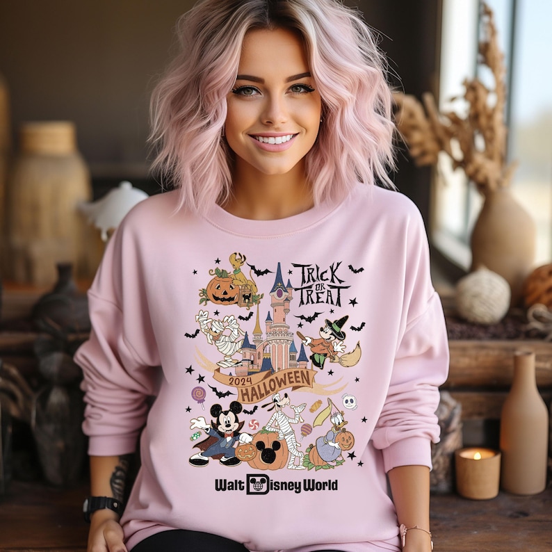 May include: Pink sweatshirt with a graphic print of Mickey Mouse, Goofy, and other Disney characters dressed in Halloween costumes. The graphic also includes a castle, pumpkins, bats, and candy. The text "Trick or Treat" and "2024 Halloween" are included in the graphic. The text "Walt Disney World" is at the bottom of the graphic.