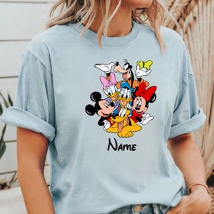Op de afbeelding: Lichtblauw T-shirt met een grafische print van Mickey Mouse, Minnie Mouse, Donald Duck, Goofy en Pluto. De tekst "NAME" is onder de personages gedrukt.