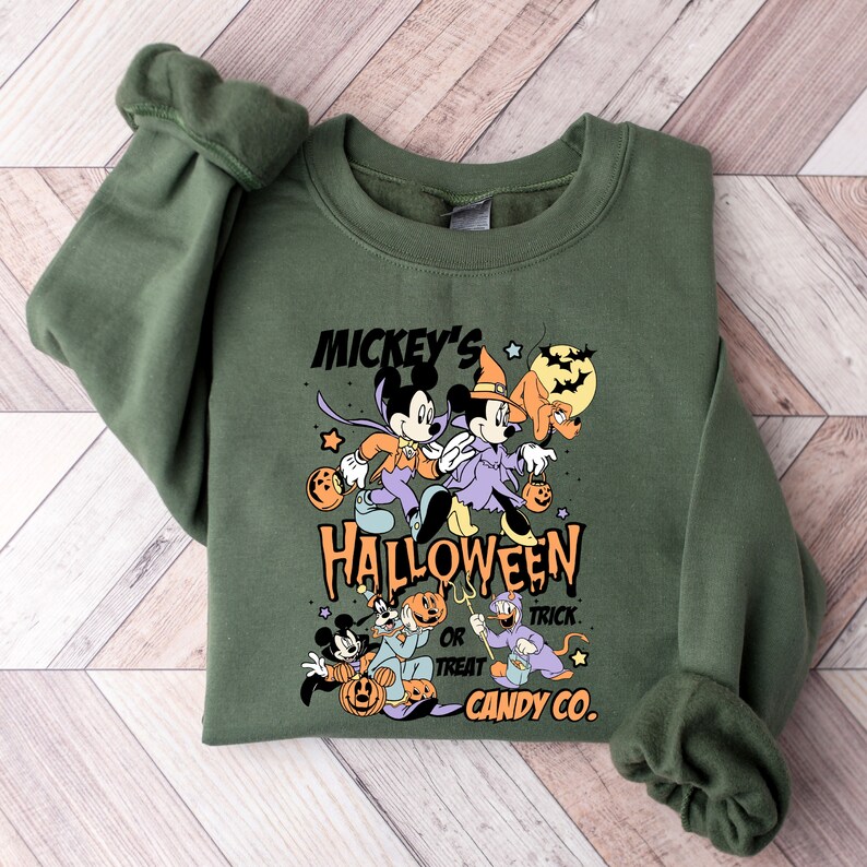 May include: Green sweatshirt with a Mickey Mouse Halloween design. The design features Mickey Mouse, Minnie Mouse, and Pluto dressed in Halloween costumes. The text on the sweatshirt reads "Mickey's Halloween or Treat Candy Co."
