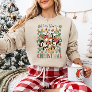 May include: Beige sweatshirt featuring a colorful graphic of Disney characters, including Mickey and Minnie Mouse, Donald Duck, and Goofy, with the text "Very Merry Christmas." The design is set against a checkered background.