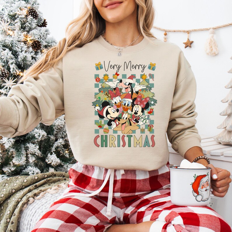 May include: Beige sweatshirt featuring a colorful graphic of Disney characters, including Mickey and Minnie Mouse, Donald Duck, and Goofy, with the text "Very Merry Christmas." The design is set against a checkered background.