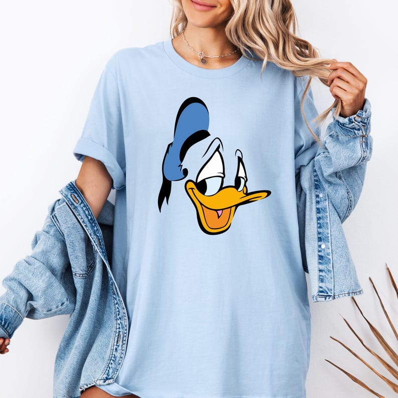 May include: Light blue t-shirt featuring a large graphic of Donald Duck's face. The cartoon character's face is in black, white, yellow, and orange. The shirt has short sleeves and a relaxed fit.