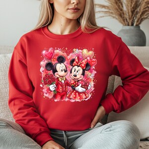 May include: Red sweatshirt featuring a colorful graphic of Mickey and Minnie Mouse in Valentine's Day attire. Mickey holds roses, and Minnie holds a heart-shaped box. Heart-shaped balloons and a pink and red background complete the design.
