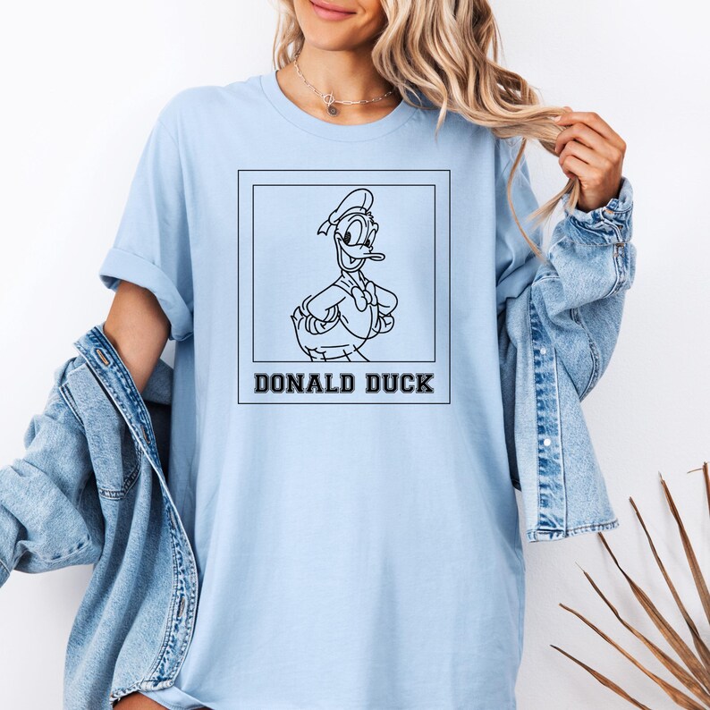May include: Light blue t-shirt featuring a black and white illustration of Donald Duck inside a square frame. The text "DONALD DUCK" is printed below the image. The shirt has long sleeves and a relaxed fit.