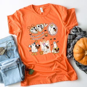 May include: Orange t-shirt with a Halloween-themed graphic. The graphic features cartoon ghosts, pumpkins, and the text "Happy Halloween." The design includes Mickey and Minnie Mouse characters in ghost costumes. The shirt is paired with blue jeans and a pumpkin.